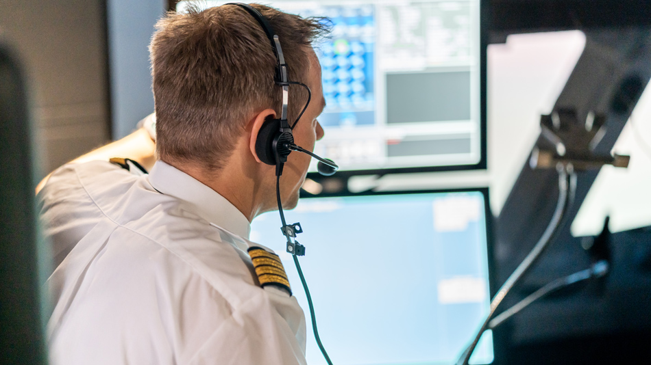 Ein Pilot als Ausbilder mit Kopfhörern auf sitzt vor Bildschirmen in einem Cockpit-Simulator und erklärt etwas