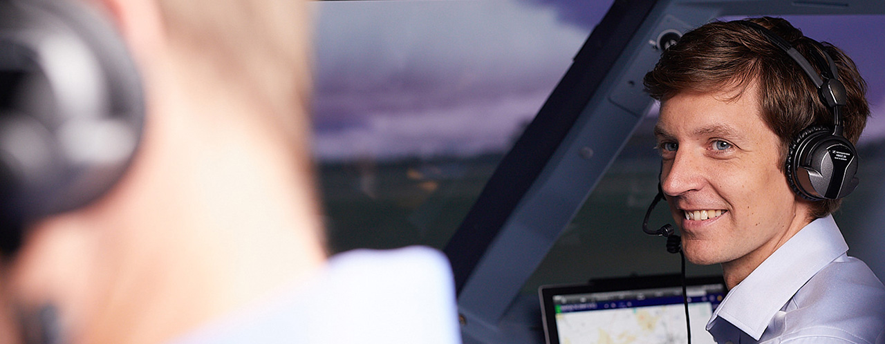 A man wearing headphones sits in an aircraft cockpit and smilingly looks at his colleague